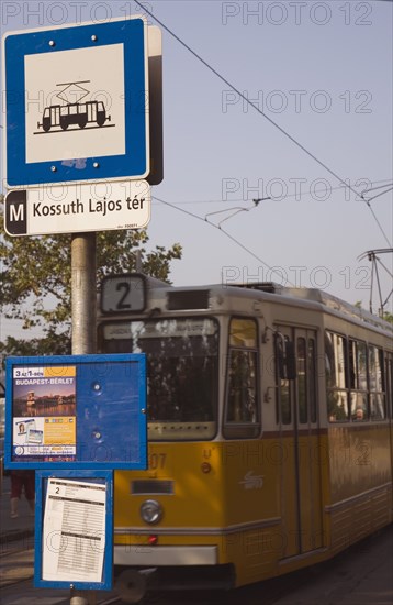 Budapest, Pest County, Hungary. Yellow tram approaching stop and signpost displaying timetable. Hungary Hungarian Europe European East Eastern Buda Pest Budapest City Urban Transport Tram Yellow Sign Timetable Eastern Europe Signs Display Posted Signage