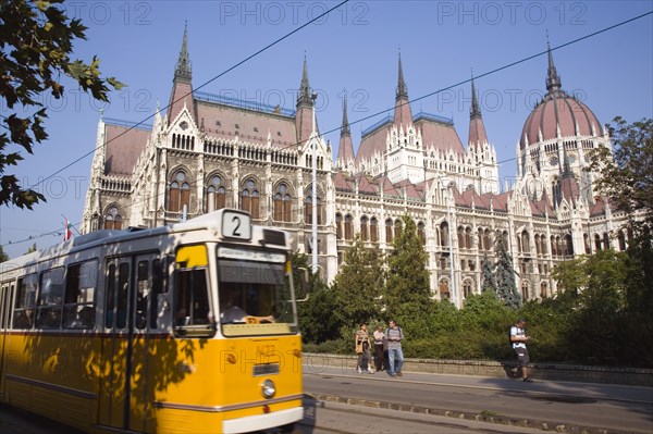 Budapest, Pest County, Hungary. Yellow tram passing the Neo-Gothic exterior facade of the Parliament Building with pedestrians on pavement beyond tram line. Hungary Hungarian Europe European East Eastern Buda Pest Budapest City Urban Transport Yellow Electric Tram Neo Gothic Parliament Building Tourists People Blue Sky Color Destination Destinations Eastern Europe Holidaymakers Parliment Tourism
