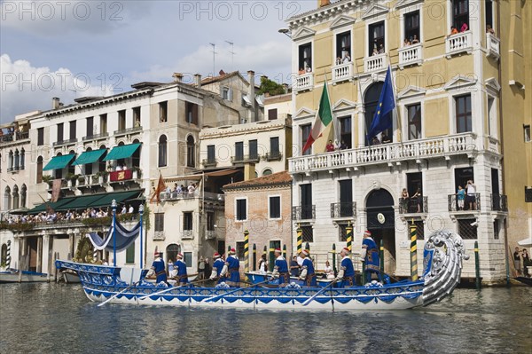Venice, Veneto, Italy. Participants in the Regata Storico historical Regatta held each September wearing traditional costume and rowing ornately decorated blue and silver gondola approaching the Rialto bridge with onlookers watching from the balconies of canalside buildings behind. Teams represent Sestiere districts of Venice in traditional races. Italy Italia Italian Venice Veneto Venezia Europe European City Regata Regatta Gondola Gondola Gondolas Gondolier Boat Architecture Exterior Water Classic Classical Clouds Cloud Sky Destination Destinations History Historic Holidaymakers Older Southern Europe Tourism Tourist