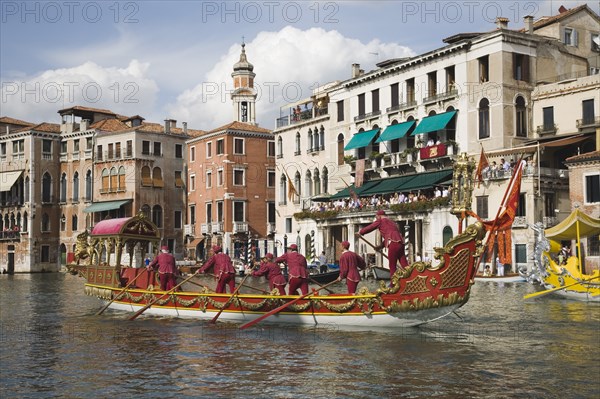 Venice, Veneto, Italy. Participants in the Regata Storico historical Regatta held each September in red and gold decorated gondola wearing traditional costume approaching the Rialto bridge with onlookers gathered on the balconies of canalside buildings behind. Teams represent Sestiere districts of Venice in traditional races. Italy Italia Italian Venice Veneto Venezia Europe European City Regata Regatta Gondola Gondola Gondolas Gondolier Boat Architecture Exterior Water Blue Classic Classical Clouds Cloud Sky Destination Destinations History Historic Holidaymakers Older Southern Europe Tourism Tourist