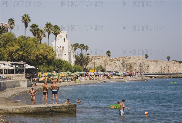Kos, Dodecanese Islands, Greece. Kos Town beach with castle walls and harbour entrance behind and Greek holidaymakers bathing on shingle beach in foreground. Greece Greek Europe European Vacation Holiday Holidays Travel Destination Tourism Ellas Hellenic Dodecanese Kos Beach Castle Walls Swimmimers Swimming Sunbtherrs Sunbathing Travel Castillo Castello Destination Destinations Ellada Sand Sandy Beaches Tourism Seaside Shore Tourist Tourists Vacation Southern Europe Sunbather