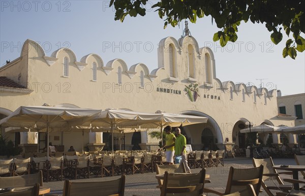 Kos, Dodecanese Islands, Greece. Kos Town Agora market in Summer sun with holidaymakers walking past near empty cafe seating. Greece Greek Europe European Vacation Holiday Holidays Travel Destination Tourism Ellas Hellenic Docecanese Kos Agora MArket Cafe Seats Tables Outside Tourists Bar Bistro Destination Destinations Ellada Restaurant Southern Europe