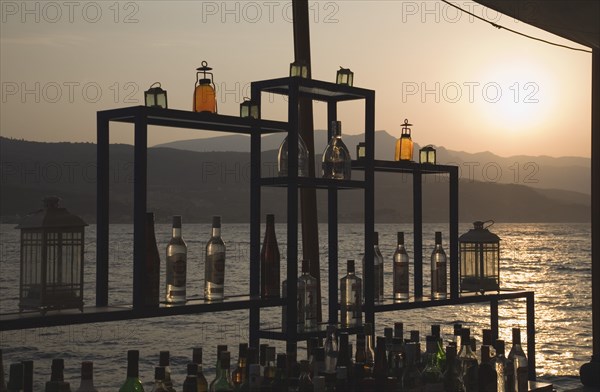 Samos, Northern Aegean, Greece. Vathy. Sun setting over water behind open air bar with the Ampelos massif beyond. Greece Greek Europe European Vacation Holiday Holidays Travel Destination Tourism Ellas Hellenic Northern Aegean Samos Island Sea Water Ampelos Massif Bar Drinks Bottles Alcohol Sun Silhouette Silhouetted Destination Destinations Ellada Solid Outline Shade Silhouette Solid Outline Shade Silhouetted Southern Europe Sundown Atmospheric
