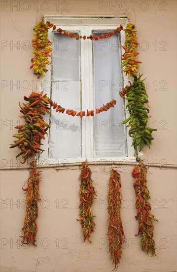 Kusadasi, Aydin Province, Turkey. Strings of red green and orange chili peppers strung up to dry in summer sunshine from the window frame of house in old town. Turkey Turkish Eurasia Eurasian Europe Asia Turkiye Aydin Province Kusadasi Chilli Chilli Chillis Chillis Chilie Chillies Pepper Peppers Capsicum Capsicums Window Drying Red Color Destination Destinations European Middle East South Eastern Europe Western Asia