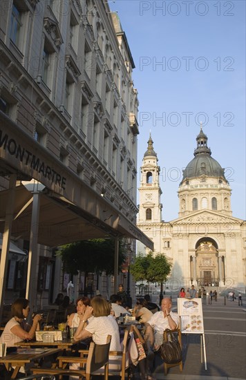 Budapest, Pest County, Hungary. Cafe with Summer tourists seated at outside tables with Saint Stephens Basilica behind. Hungary Hungarian Europe European East Eastern Buda Pest Budapest City Cafe Restaurant Tables Outside Al Fresco Sat People Tourists St Saint Stephen Stephens Basilica Church Bar Bistro Blue Destination Destinations Eastern Europe Holidaymakers Religion Religious Tourism
