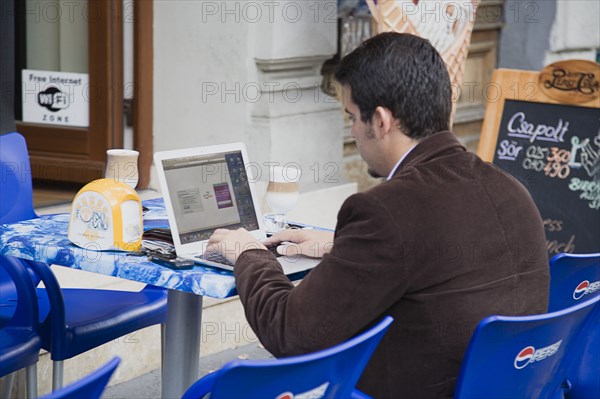 Budapest, Pest County, Hungary. Hungarian man has coffee at outside table while using laptop online with wifi access on Andrassy Ut In Pest. Window sign advertises free internet access and wifi zone. Hungary Hungarian Europe European East Eastern Buda Pest Budapest City Markets Food Drink Cafe Restaurant Coffee Internet Wifi Computer Laptop PC Personal Man Using Bar Bistro Eastern Europe Male Men Guy One individual Solo Lone Solitary Signs Display Posted Signage