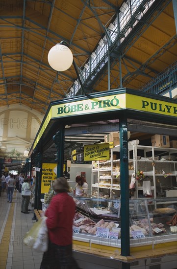 Budapest, Pest County, Hungary. Stall selling Hungarian sausages hams bacon and other meats at Nagy Vasarcsarnok the indoor Central Market. Woman carrying shopping bags walks past in foreground. Hungary Hungarian Europe European East Eastern Buda Pest Budapest City Nagy Vasarcsarnok Central Market Markets Indoor Inside Interior Stall Shop Store Meat Meats Cooked Raw Sausage Sausages Ham bacon Woman Carrying Shopping Bags Destination Destinations Eastern Europe Female Women Girl Lady Shops Shoppers Mall Retail Buy Buying Market Markets