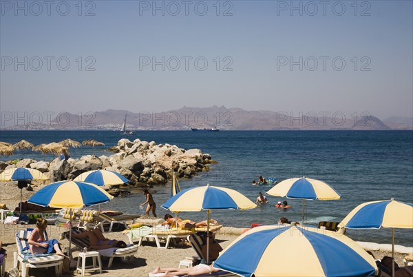 Kos, Dodecanese Islands, Greece. Tigaki Beach. Sunbathers on loungers with blue and yellow striped beach parasols with others swimming in sea and the Turkish coast of Bodrum behind. Greece Greek Europe European Vacation Holiday Holidays Travel Destination Tourism Ellas Hellenic Docecanese Kos Tigaki Beach Sun Loungers Sunbathers Sunbathing Tourist Tourists Umbrellas Parasols Sea Color Destination Destinations Ellada Holidaymakers Sand Sandy Beaches Tourism Seaside Shore Tourist Tourists Vacation Sightseeing Southern Europe Water