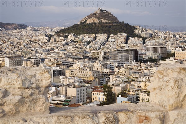 Athens, Attica, Greece. Mount Lycabettus rising in central Athens with densely populated city below part framed by wall in foreground. Greece Greek Europe European Vacation Holiday Holidays Travel Destination Tourism Ellas Hellenic Attica Athens City View Cityscape Aerial Elevated Houses Housing Mount Lycabettus Atenas Athenes Destination Destinations Ellada Southern Europe