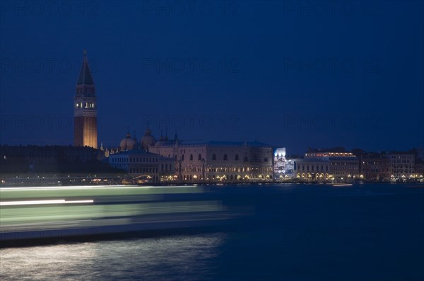 Venice, Veneto, Italy. Giudecca island Early evening view towards illuminated Campanile of St. Mark with vaporetto water bus to Murano and Burano in blurred motion in foreground with light trails reflected in water of the canal. Italy Italia Italian Venice Veneto Venezia Europe European City Giudecca Island St saint Mark Marks Campanile Vaporetto Water Bus Blur Blurred Movement Streak Streaks Streaked Light Lights Transport Night Illuminated Destination Destinations Nite Religion Southern Europe Warm Light