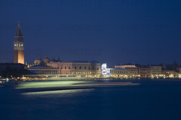 Venice, Veneto, Italy. Giudecca island Early evening view towards illuminated Campanile of St. Mark with vaporetto water bus to Murano and Burano in blurred motion in foreground with light trails reflected in water of the canal. Italy Italia Italian Venice Veneto Venezia Europe European City Illuminated Night Transport Transport Light Streaked Streaks Streak Movement Blurred Blur Bus Water Vaporetto Campanile St Saint Mark Marks Destination Destinations Nite Religion Southern Europe Warm Light