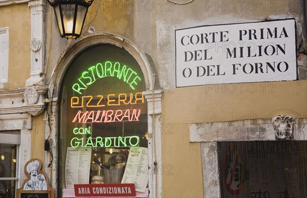 Venice, Veneto, Italy. Centro Storico Pizzeria window in old city wall with neon advertising menus and sign for air conditioning. Painted street sign on wall at side above doorway. Italy Italia Italian Venice Veneto Venezia Europe European City Cenro Stroico Pizzeria Window Neon Sign Signs Colour Color Colors Colours Colorful Colourful Multi Multiple Facade Exterior Architecture Signs Display Posted Signage Southern Europe