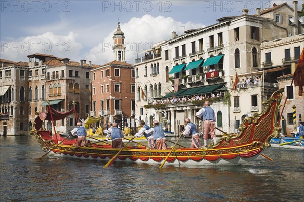 Venice, Veneto, Italy. Participants in the Regata Storico historical Regatta held each September rowing red and gold decorated gondola and wearing traditional costume approaching the Rialto bridge with onlookers gathered on the balconies of canalside buildings behind. Teams represent Sestiere districts of Venice in traditional races. Italy Italia Italian Venice Veneto Venezia Europe European City Regata Regatta Gondola Gondola Gondolas Gondolier Boat Architecture Exterior Water Blue Classic Classical Clouds Cloud Sky Destination Destinations History Historic Holidaymakers Older Southern Europe Tourism Tourist