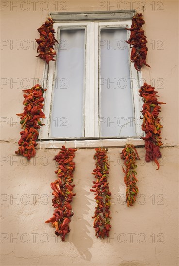 Kusadasi, Aydin Province, Turkey. Strings of red and orange chili peppers hung up to dry in summer sunshine from window frame of house in the old town. Turkey Turkish Eurasia Eurasian Europe Asia Turkiye Aydin Province Kusadasi Chilli Chilli Chillis Chillis Chilie Chillies Pepper Peppers Capsicum Capsicums Window Drying Red Color Destination Destinations European Middle East South Eastern Europe Western Asia