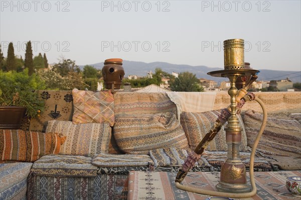 Selcuk, Izmir Province, Turkey. Ephesus. View from rooftop cafe with water pipe on table in foreground and traditional kilim textiles. Turkey Turkish Eurasia Eurasian Europe Asia Turkiye Izmir Province Selcuk Ephesus Cafe Rooftop Waterpipe Water Pipe Smoke Smoking Kilim Textiles Bar Bistro Classic Classical Cloth Destination Destinations European Historical History Historic Middle East Older Restaurant South Eastern Europe Western Asia
