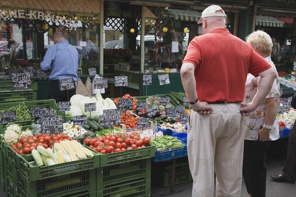 Vienna, Austria. The Naschmarkt. Tourists looking at display on fresh produce stall in front of glass fronted shop front including aubergines tomatoes sweetcorn and cauliflower. Austrian Destination Destinations European Holidaymakers Osterreich Store Tourism Viena Western Europe Wien