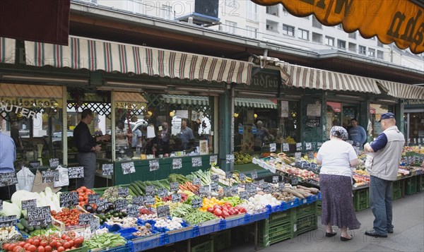 Vienna, Austria. The Naschmarkt. Customers at fresh produce stall with display including peppers leeks tomatoes and mushrooms in foreground of glass shopfronts. Austria Austrian Republic Vienna Viennese Wien Europe European City Capital Naschmarkt Market Display Fresh Food Fruit Vegetables Veg Vegetable Stall Shop Store Destination Destinations Osterreich Viena Western Europe