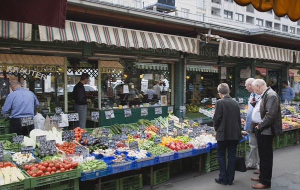 Vienna, Austria. The Naschmarkt. Customers at fresh produce stall looking at display that includes tomatoes aubergines leeks courgettes and mushrooms. Austria Austrian Republic Vienna Viennese Wien Europe European City Capital Naschmarkt Market Display Fresh Food Fruit Vegetables Veg Vegetable Stall Shop Store Destination Destinations Osterreich Viena Western Europe