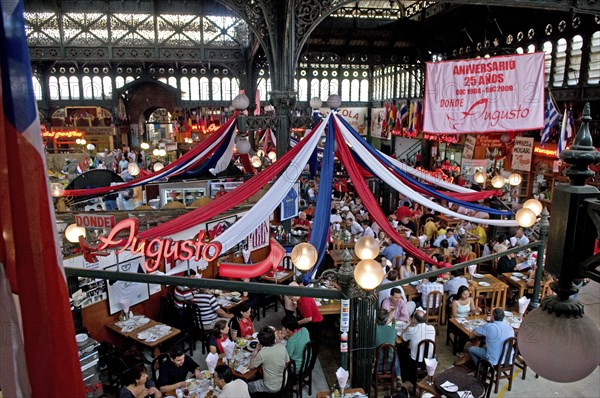 Santiago, Chile. Seafood restaurant section of Central Market with diners eating at tables under red white and blue hanging decorations. The ironwork used in the structure of the building which was completed in 1872 was made in England. Chile Chilean South America American Hispanic Santiago City Cityscape Urban Travel Destination Vacation Holiday Mercado Central Market Retaurant Cafe Seafood Fish View Over Diners People Crowded Tables Eating Food Drink Bunting Streamers Bar Bistro Destination Destinations Great Britain Latin America Latino Northern Europe South America Southern UK United Kingdom