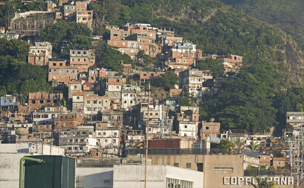 Rio de Janeiro, Brazil. Favela or slum on hillside above Copacabana neighbourhood greenery and COPACABANA in large letters on building in foreground. Brazil Brasil Brazilian Brasilian South America Latin Latino American City Urban Architecture Houses Housing Homes Copacabana Neighborhood Neighbourhood Favela Favelas Slum Slums Church Rio de Janeiro Destination Destinations Latin America Shanty South America Southern