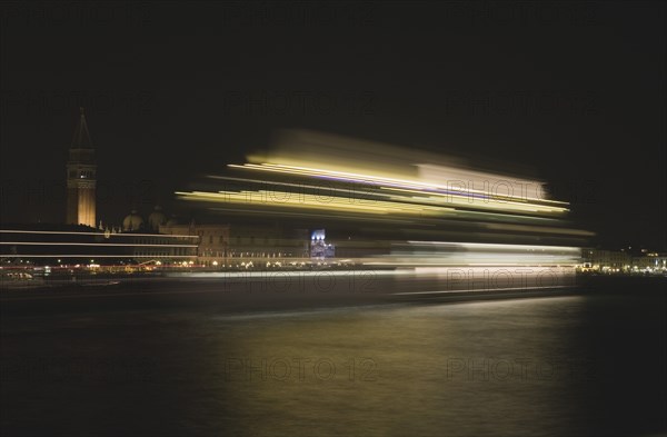 Venice, Veneto, Italy. Giudecca island Evening view towards illuminated Campanile of St. Mark with vaporetto water bus to Murano and Burano in blurred motion in foreground with light trails reflected in water of the canal. Italy Italia Italian Venice Veneto Venezia Europe European City Marks Mark Saint St Campanile Vaporetto Water Water Blur Blurred Movement Streak Streaks Streaked Light Lights Illuminated Destination Destinations Religion Southern Europe Warm Light