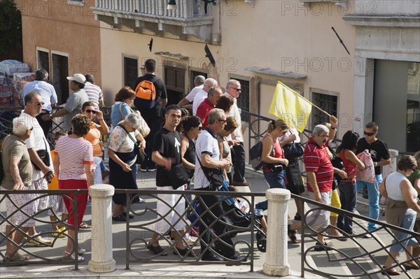 Venice, Veneto, Italy. Tour group of tourists crossing bridge in late summer led by guide holding flag above to show direction. Italy Italia Italian Venice Veneto Venezia Europe European City Canal Bridge Tourist Tourists People Crowd Group Crossing Tour Guide Flag Destination Destinations Holidaymakers Sightseeing Southern Europe Tourism