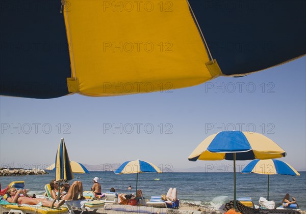 Kos, Dodecanese Islands, Greece. Sunbathers on loungers and blue and yellow striped beach parasols on beach outside Kos Town. Greece Greek Europe European Vacation Holiday Holidays Travel Destination Tourism Ellas Hellenic Docecanese Kos Beach Sun Loungers Sunbathers Sunbathing Tourists Umbrellas Parasols Sea Yellow Blue Color Destination Destinations Ellada Holidaymakers Sand Sandy Beaches Tourism Seaside Shore Tourist Tourists Vacation Southern Europe Water