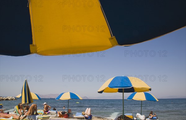 Kos, Dodecanese Islands, Greece. People sunbathing on loungers some beneath yellow and blue striped parasols on beach outside Kos Town. Greece Greek Europe European Vacation Holiday Holidays Travel Destination Tourism Ellas Hellenic Docecanese Beach Beach Sun Loungers Sunbathers Sunbathing Tourists Umbrellas Parasols Sea Yellow Blue Color Destination Destinations Ellada Holidaymakers Sand Sandy Beaches Tourism Seaside Shore Tourist Tourists Vacation Southern Europe Water