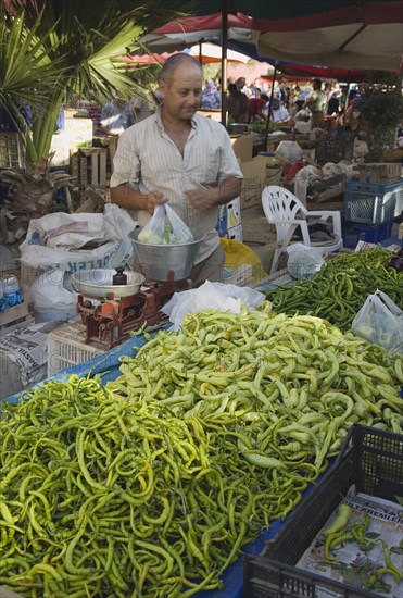Kusadasi, Aydin Province, Turkey. Turkish stallholder selling green chilis under shade in town weekly produce market using set of scales to weigh bag of goods. Asian Color Destination Destinations European Middle East One individual Solo Lone Solitary South Eastern Europe Turkiye Western Asia