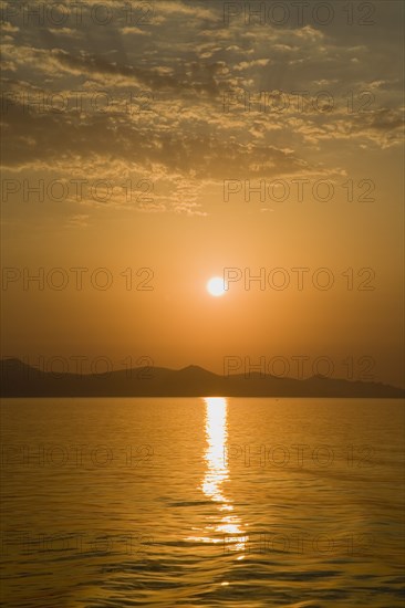 Samos, Northern Aegean, Greece. Vathy. Sunset over Samos seen from ferry between Samos and Athens as it leaves Vathy Samos with gold and orange sky reflected in water and silhouetted landscape. Greece Greek Europe European Vacation Holiday Holidays Travel Destination Tourism Ellas Hellenic Nortehrn Aegean Samos Island Vathy Sunet Sunt Orange Sky Sea Water Reflected Reflection Ripples Atenas Athenes Blue Color Destination Destinations Ellada Reflexion Scenic Solid Outline Shade Silhouette Southern Europe Sundown Atmospheric