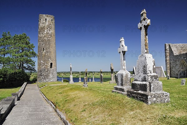 Clonmacnoise, County Offaly, Ireland. Monastery Round tower with row of crosses and River Shannon in the background. Ireland Irish Eire Erin Europe European Couty Offaly Clonmacnoise Monastery Round Tower Cross Crosses Crucifix Crucifixes Blue Sky River Shannon Water Celtic Color Destination Destinations Gray History Historic Northern Europe Poblacht na hEireann Religion Religious Republic Colour Grey