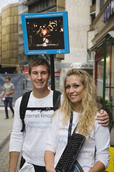 Budapest, Pest County, Hungary. Attractive young student couple promoting mobile internet access near Saint Stephens Basilica. Hungary Hungarian Europe European East Eastern Buda Pest Budapest City Couple Young Boy Girl man Woman PR Promo Promote Promoting Internet Access Server Cell Cellular Eastern Europe Female Women Girl Lady Immature Male Men Guy
