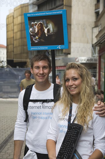 Budapest, Pest County, Hungary. Attractive young student couple promoting mobile internet access near Saint Stephens Basilica. Hungary Hungarian Europe European East Eastern Buda Pest Budapest City Couple Young Girl Girl man Woman PR Promo Promote Promoting Internet Access Server Cell Cellular Eastern Europe Female Women Girl Lady Immature Male Men Guy