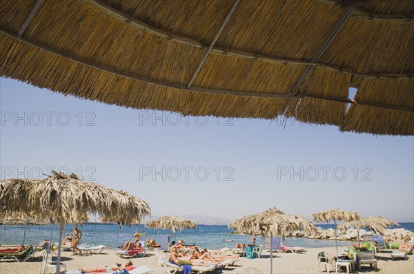 Kos, Dodecanese Islands, Greece. People sunbathing on beach outside Kos Town part framed by thatched shade. Greece Greek Europe European Vacation Holiday Holidays Travel Destination Tourism Ellas Hellenic Docecanese Kos Beach Loungers Loungers Sunbathers Sunbathing Tourists Parasols Parasols Sea Blue Destination Destinations Ellada Holidaymakers Sand Sandy Beaches Tourism Seaside Shore Tourist Tourists Vacation Southern Europe Water