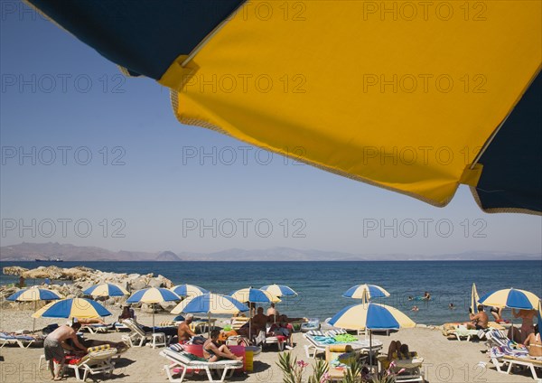 Kos, Dodecanese Islands, Greece. People sunbathing on beach outside Kos Town with sun loungers blue and yellow striped parasols and people in shallow water of rock enclosed bay beyond. Part framed by underside of parasol in foreground. Greece Greek Europe European Vacation Holiday Holidays Travel Destination Tourism Ellas Hellenic Docecanese Kos Beach Sun Sunbathers Sunbathing Loungers Tourists Parasols Sea Umbrellas Blue Yellow Color Destination Destinations Ellada Holidaymakers Sand Sandy Beaches Tourism Seaside Shore Tourist Tourists Vacation Southern Europe