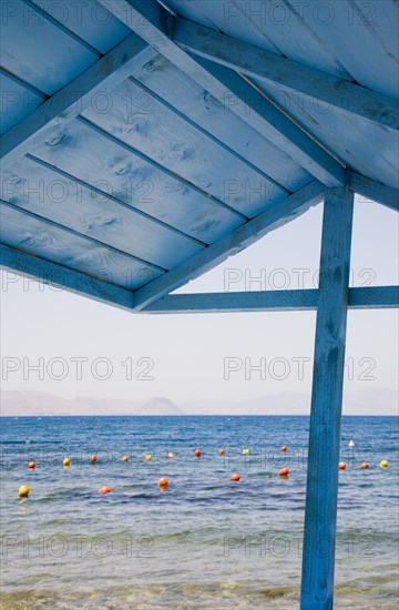 Kos, Dodecanese, Greece. View over red and yellow floats in water part framed by roof of blue painted wooden structure in foreground. Greece Greek Europe European Vacation Holiday Holidays Travel Destination Tourism Ellas Hellenic Docecanese Kos Beach Sea Blue Wooden Wood Shelter Buoys Color Destination Destinations Ellada Sand Sandy Beaches Tourism Seaside Shore Tourist Tourists Vacation Southern Europe