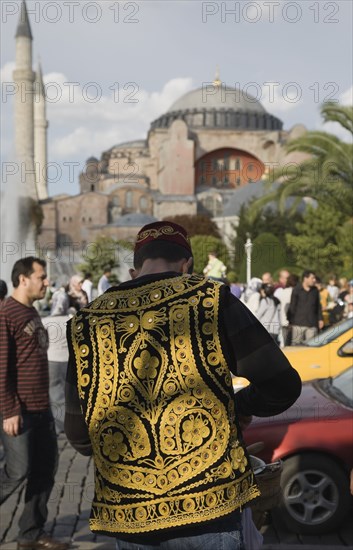 Istanbul, Turkey. Sultanahmet Man wearing embroidered waistcoat and cap standing at food stall in front of Hagia Sophia part seen beyond. Turkey Turkish Istanbul Constantinople Stamboul Stambul City Europe European Asia Asian East West Urban Destination Travel Tourism Sultanahmet Hagia Sophia Mosque Man Traditional Dress Costuime Embroidered Waistcoat Vest Middle East South Eastern Europe Turkish Turkiye Western Asia