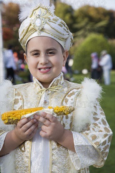 Istanbul, Turkey. Sultanahmet. Young boy eating sweetcorn cooked on the cob wearing traditional Turkish ceremonial attire standing in front of the Blue Mosque on the day of his circumcision. Turkey Turkish Istanbul Constantinople Stamboul Stambul City Europe European Asia Asian East West Urban Destination Travel Tourism Young Boy Draditional Dress Costume eating Corn Cob Classic Classical Cultural Cultures Destination Destinations Historical Immature Kids Middle East Older One individual Solo Lone Solitary Order Fellowship Guild Club South Eastern Europe Turkiye Western Asia