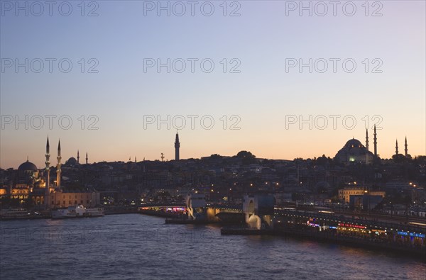 Istanbul, Turkey. Sultanahmet. The Golden Horn. The New Mosque or Yeni Camii at left the Galata Bridge and Suleymaniye Mosque at right with lights and advertising illuminated at dusk Turkey Turkish Istanbul Constantinople Stamboul Stambul City Europe European Asia Asian East West Urban Skyline Destination Travel Tourism Sultanahmet Golden Horn New Mosque Yeni Camii Galata Bridge Suleymaniye Dusk Sunset Night Illuminated Skyline Minarets Water Citiscape Building Buildings Urban Architecture Destination Destinations Middle East Nightfall Twilight Evenfall Crespuscle Crespuscule Gloam Gloaming Nite Religion Religious South Eastern Europe Sundown Atmospheric Turkiye Western Asia