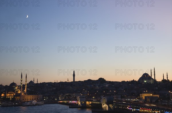Istanbul, Turkey. Sultanahmet. The Golden Horn. The New Mosque or Yeni Camii at left the Galata Bridge and Suleymaniye Mosque at right. Buildings illuminated at dusk and crescent moon in purple blue sky above. Turkey Turkish Istanbul Constantinople Stamboul Stambul City Europe European Asia Asian East West Urban Skyline Destination Travel Tourism Sultanahmet Golden Horn New Mosque Yeni Camii Galata Bridge Suleymaniye Dusk Sunset Night Illuminated Skyline Minarets Water Crescent Moon Citiscape Building Buildings Urban Architecture Destination Destinations Middle East Nightfall Twilight Evenfall Crespuscle Crespuscule Gloam Gloaming Nite Religion Religious South Eastern Europe Sundown Atmospheric Turkiye Western Asia