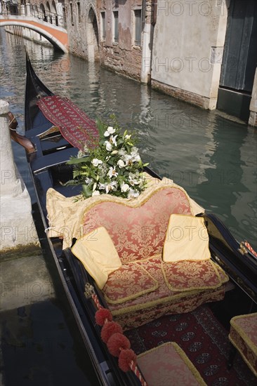 Venice, Veneto, Italy. Gondola prepared for wedding with red and gold brocade seating and flowers moored at canal jetty. Italy Italia Italian Venice Veneto Venezia Europe European City Water Canal Bridge Gondola Boat Red Gold Cushions Seats Moored Jetty Flowers Destination Destinations Marriage Marrying Espousing Hymeneals Nuptials Religion Southern Europe
