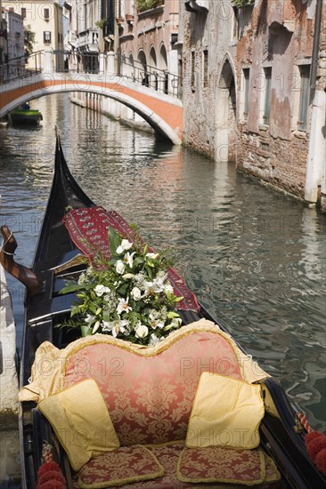 Venice, Veneto, Italy. Gondola prepared for wedding with flowers and red and gold brocade seating moored at canal jetty. Italy Italia Italian Venice Veneto Venezia Europe European City Water Canal Bridge Gondola Boat Red Gold Cushions Seats Moored Jetty Flowers Destination Destinations Marriage Marrying Espousing Hymeneals Nuptials Religion Southern Europe