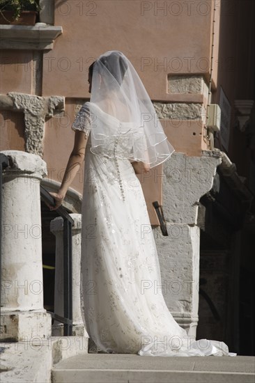 Venice, Veneto, Italy. Bride in white wedding dress and veil standing on canal bridge in late summer sun back to camera. Italy Italia Italian Venice Veneto Venezia Europe European City Wedding Wed Marriage Bride Dress White Lace Veil Bridge Canal Pose Posing Color Marriage Marrying Espousing Hymeneals Nuptials One individual Solo Lone Solitary Religion Southern Europe