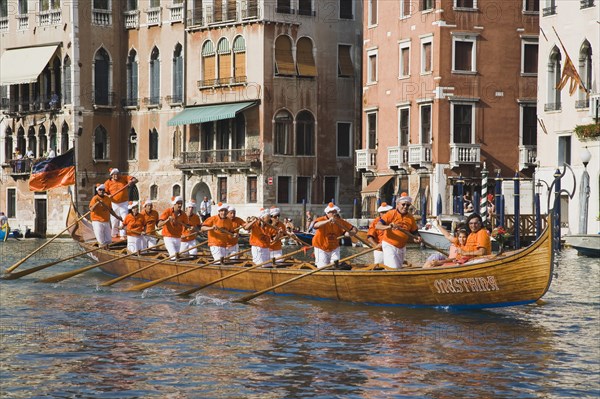 Venice, Veneto, Italy. Regatta Storico historical annual regatta. Team of rowers wearing orange and white costume passing canalside buildings with onlookers watching from balconies. Italy Italia Italian Venice Veneto Venezia Europe European City Regata Regatta Gondola Gondola Gondolas Gondolier Boat Architecture Exterior Water Destination Destinations History Historic Holidaymakers Southern Europe Tourism Tourist