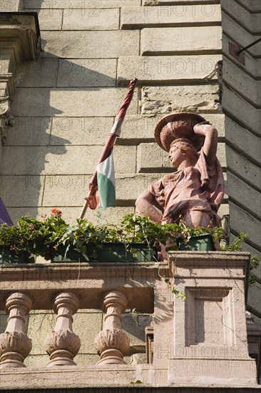 Budapest, Pest County, Hungary. Art Nouveau exterior facade with rolled Hungarian flag flower pots and statue on stone balcony. Hungary Hungarian Europe European East Eastern Buda Pest Budapest City Architecture Art Nouveau Facade Detail Stone Balcony Statue Flowers Pots Destination Destinations Eastern Europe