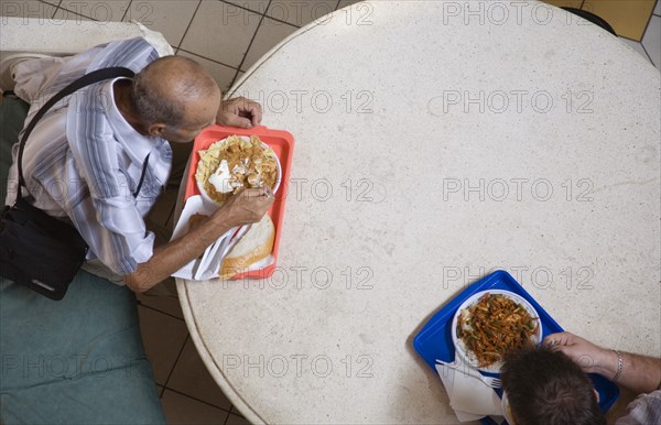 Budapest, Pest County, Hungary. Looking down on two men having lunchtime meal seated at circular table in Nagy Vasarcsarnok the Central Market. Hungary Hungarian Europe European East Eastern Buda Pest Budapest City Nagy Vasarcsarnok Central Market Food Eating lunch Lunchtime Meal Men Table Seated Aerial Trays 2 Eastern Europe Male Man Guy