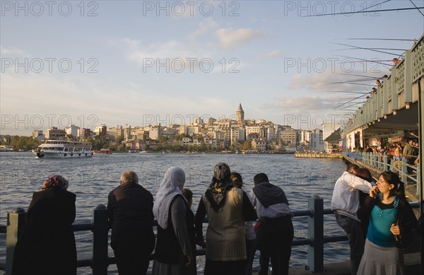 Istanbul, Turkey. Sultanahmet. Crowd leaning on railings beside the Bosphorus admiring view to Galata tower with rods of men fishing from Galata bridge extended out above. Turkey Turkish Istanbul Constantinople Stamboul Stambul City Europe European Asia Asian East West Urban Destination Travel Tourism Sultanahmet Bosphorous Galata Bridge People Crowd Fishing Rods Men Destination Destinations Male Man Guy Middle East South Eastern Europe Turkiye Western Asia