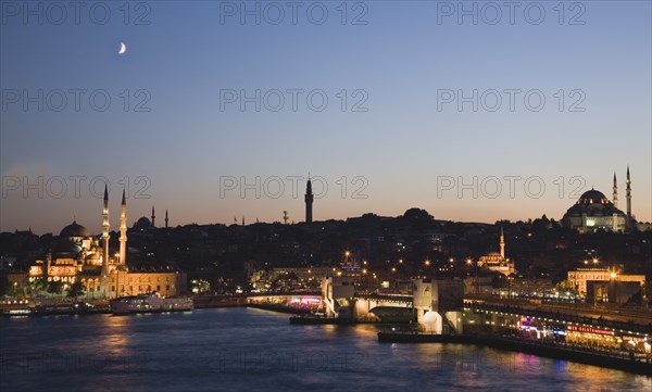 Istanbul, Turkey. Sultanahmet. The Golden Horn. The New Mosque or Yeni Camii at left the Galata Bridge and Suleymaniye Mosque at right with buildings and restaurants illuminated at dusk and crescent moon in blue purple sky above. Turkey Turkish Istanbul Constantinople Stamboul Stambul City Europe European Asia Asian East West Urban Skyline Destination Travel Tourism Sultanahmet Golden Horn New Mosque Yeni Camii Galata Bridge Suleymaniye Dusk Sunset Night Illuminated Skyline Minarets Water Crescent Moon Citiscape Building Buildings Urban Architecture Destination Destinations Middle East Nightfall Twilight Evenfall Crespuscle Crespuscule Gloam Gloaming Nite South Eastern Europe Sundown Atmospheric Turkiye Western Asia