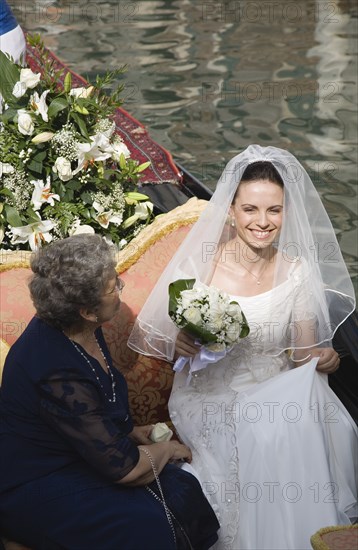 Venice, Veneto, Italy. Smiling bride in wedding dress and veil carrying bouquet of white roses seated beside mother on gondola prepared for wedding trip on canal in late summer sunshine. European Happy Italia Italian Marriage Marrying Espousing Hymeneals Nuptials Mum Religion Southern Europe