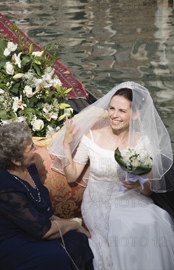 Venice, Veneto, Italy. Smiling bride in wedding dress and veil carrying bouquet seated beside mother on gondola prepared for wedding trip on canal in late summer sunshine. Italy Italia Italian Venice Veneto Venezia Europe European City Wed Wedding Marriage Dress Bride White Lace Veil Canal Gondola Smiling Happy Flowers Bouquet Contented Marriage Marrying Espousing Hymeneals Nuptials Mum Religion Southern Europe